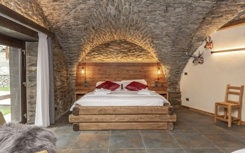 Rustic room with a bed, wooden chair, stone wall, deer heads, and a doorway leading to a grassy area outside.