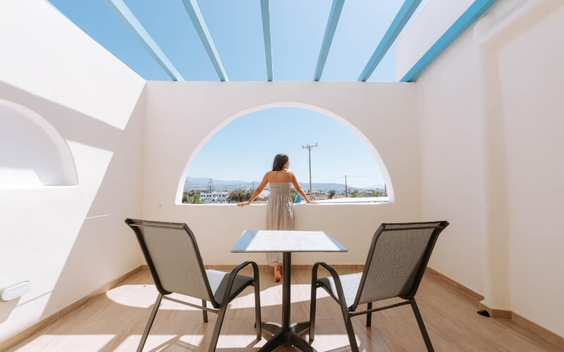 Donna in abito bianco su un balcone, guarda un paesaggio sereno attraverso una grande finestra ad arco.