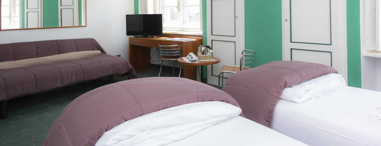 Cozy hotel room with two beds, a couch, a desk with a TV, and a large window, decorated in white and green.