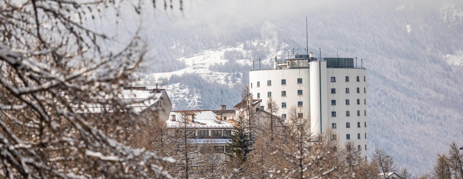 Scena invernale con un edificio cilindrico bianco centrale, alberi e montagne innevate sullo sfondo.