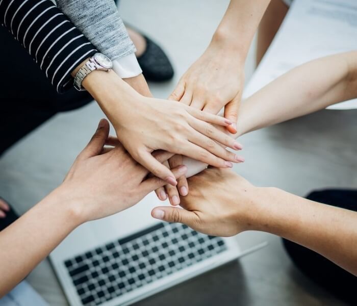 Cinque persone formano un cerchio con le mani unite sopra una tastiera di laptop sfocata su una superficie grigia.