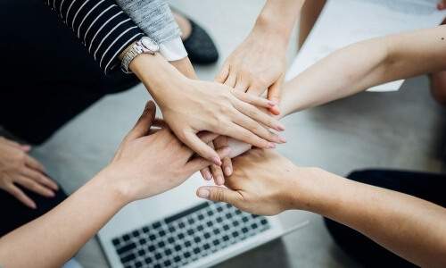 A group of hands stacked together, with a laptop and paper in the background, symbolizing teamwork and support.