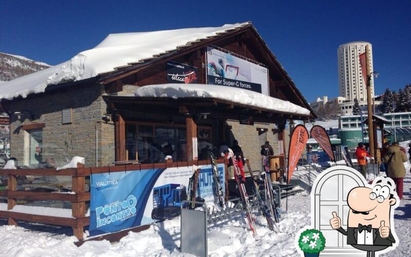 Snow-covered building with sloping roof, signs, and people in winter attire near stacked skis and snowboards.