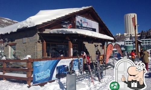 Rustic wooden building with snow, Val Thorens banner, ski equipment outside, clear sky, and mountains in the background.