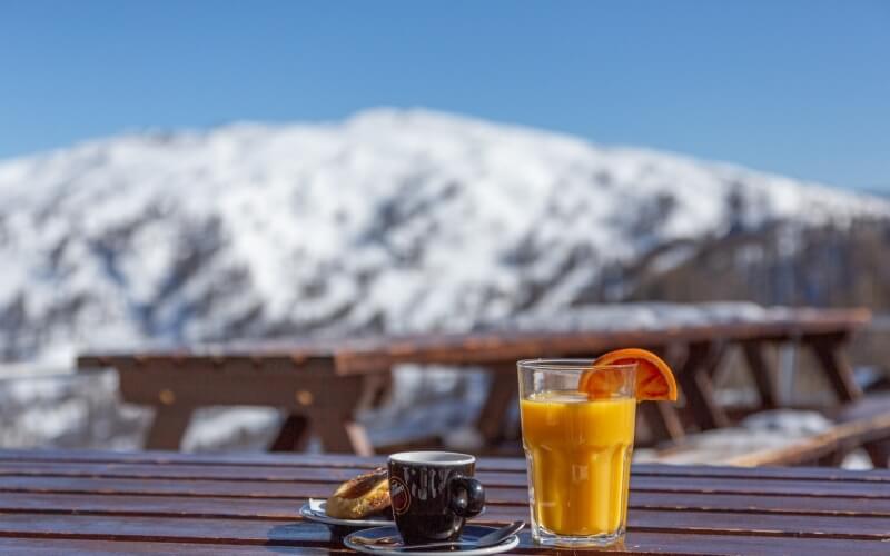 Wooden table with coffee, orange juice, and pastry, set against a snowy mountain backdrop on a sunny day.