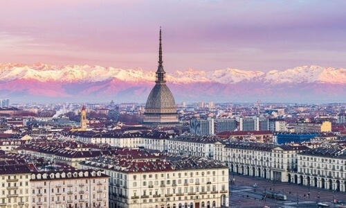 Serene cityscape of Turin, Italy, featuring beige buildings, a church steeple, and a mountain range under a pink sky.