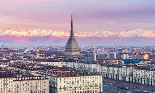 Un paesaggio urbano affollato con un edificio centrale distintivo e montagne innevate sullo sfondo.