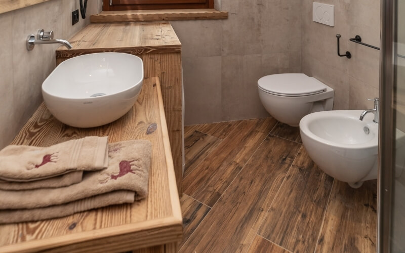 A clean bathroom featuring a light wood vanity, oval sink, beige towels, toilet, and large gray tile walls.