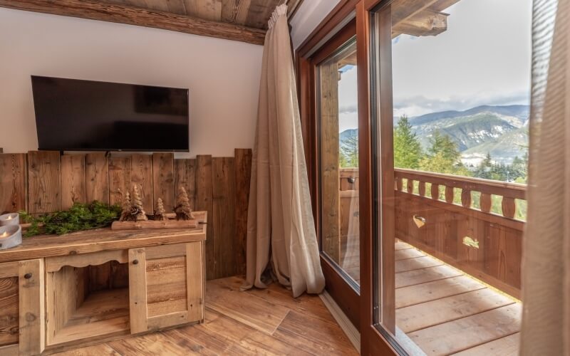 Cozy room with a TV and wooden deck, showcasing serene mountains and trees under a light gray sky.