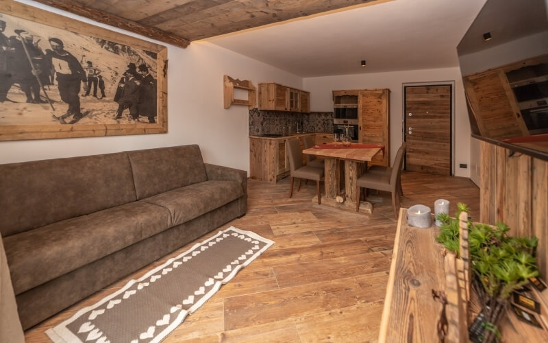 Rustic living room and kitchen with a brown couch, wooden table, white walls, and a large painting of people with rifles.