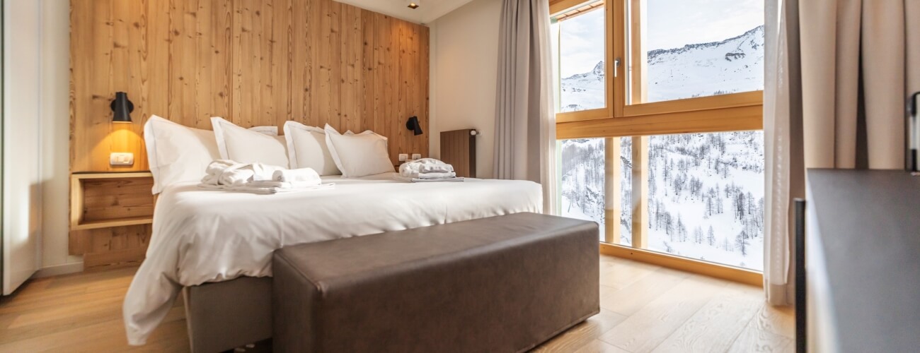 Serene bedroom with mountain views, minimalist decor, wooden accent wall, white linens, purple ottoman, and gray curtains.