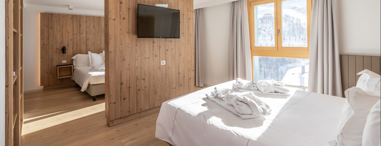 A room with wood paneling, two white beds, a large window with gray curtains, and an open doorway to another identical room.