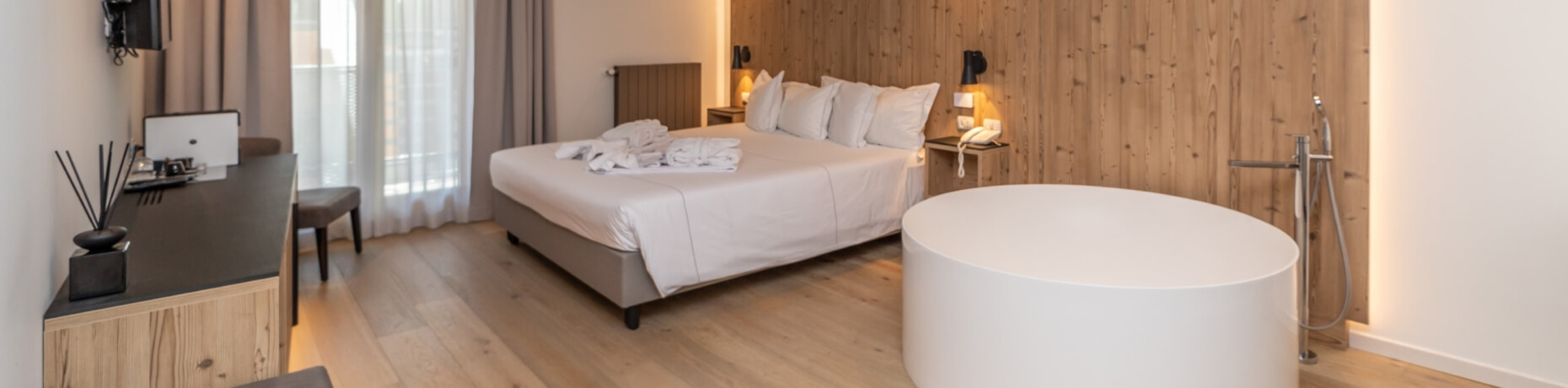 Modern hotel room with a king-sized bed, desk, bathtub, and natural light, featuring minimalistic decor and clean lines.