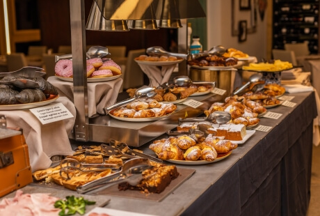 A buffet table with pastries in warming trays, brown boxes, and white cards, set in a well-lit room with framed pictures.