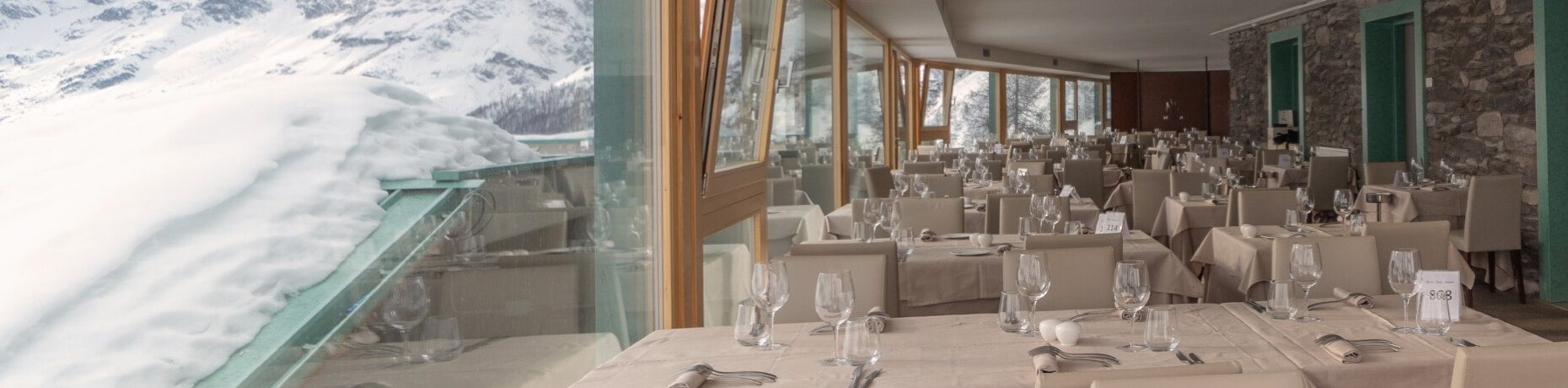 Area pranzo elegante con vista su montagne innevate, tavoli decorati con tovaglie beige e un muro di pietra caldo.