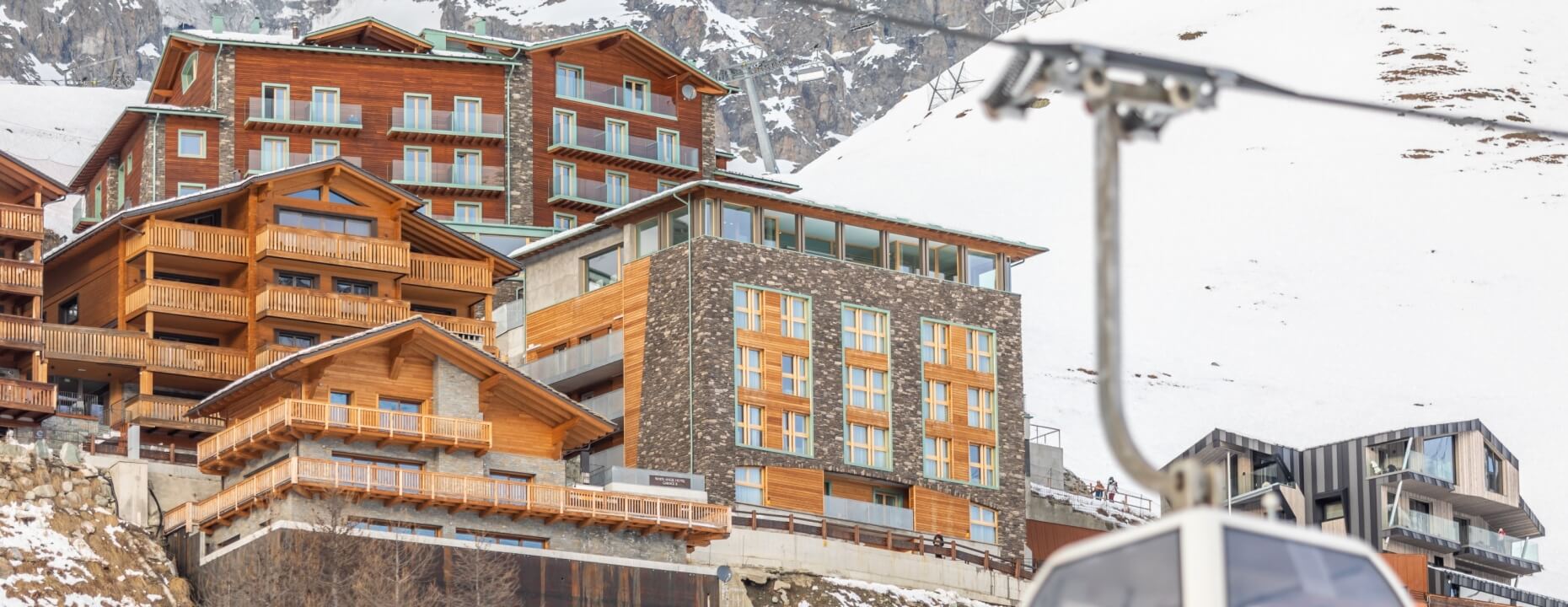 A winter scene of a wooden and stone complex at the base of a snowy mountain, with a ski lift in the foreground.