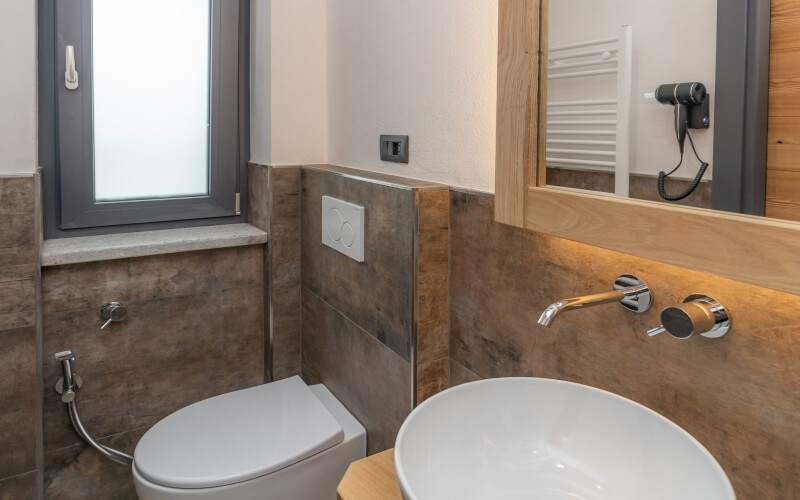 Compact bathroom with brown tiled walls, white sink, mirror with light, and window, exuding cleanliness and minimalism.