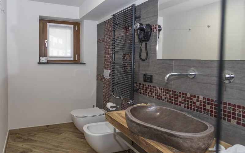 Modern bathroom featuring a sand-colored oval sink, red tiled wall, large mirror, white toilet, and a window with natural light.