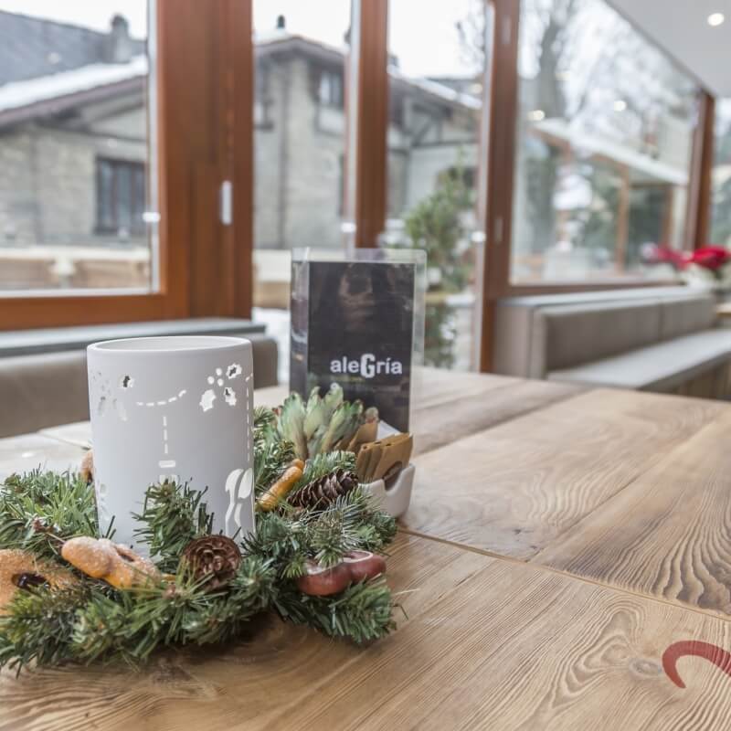 Festive table setting with a candle centerpiece, "aleGria" sign, and large windows showing a cozy residential view.