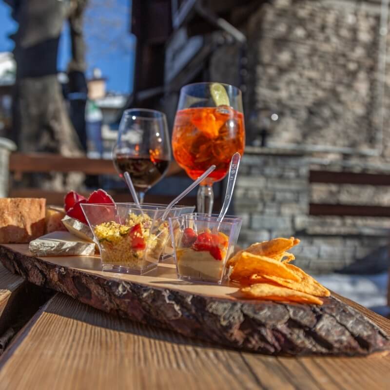 Wooden serving tray with drinks and food, set outdoors against a stone wall and trees, evoking casual dining atmosphere.
