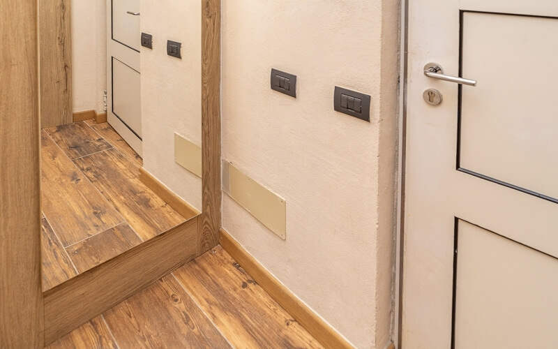 White hallway with wooden floor, large angled mirror, light switches on walls, and a white door with a silver handle.