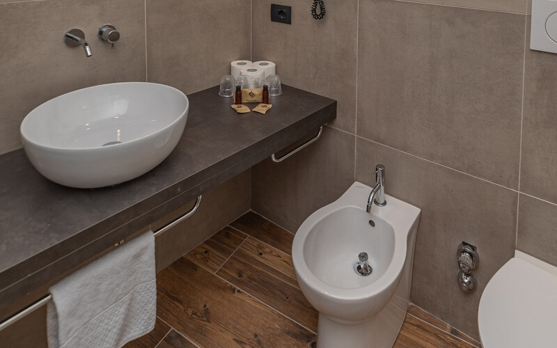 Modern bathroom with gray countertop, white sink, bidet, towel rack, and dark wood floor, featuring light-brown tiled walls.