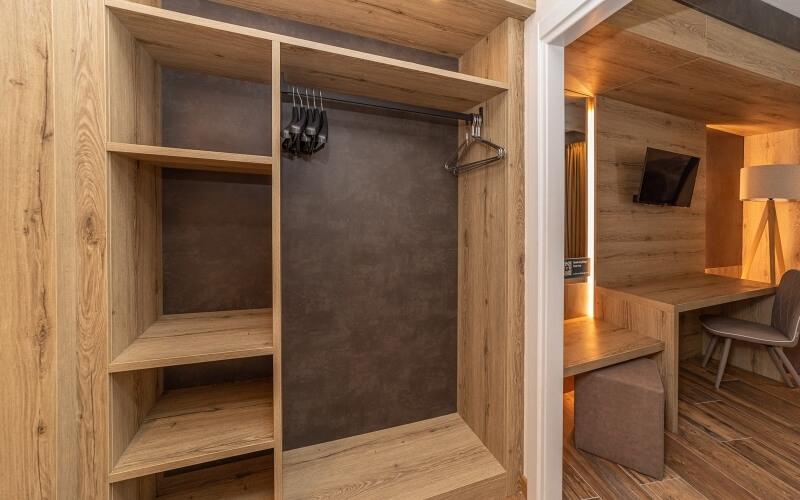 Modern hotel room featuring a large wooden closet, desk area, gray chair, lamp, TV, and small ottoman.