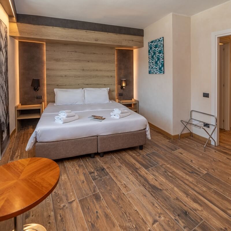 Hotel room featuring a large bed, nightstands, lamps, a brown ottoman, and a round wooden table, with white walls and wooden floor.
