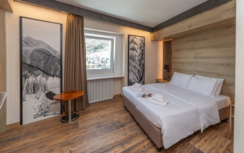 Serene hotel room with white decor, wooden accents, a window with a radiator, and a double bed beside a small table and lamp.
