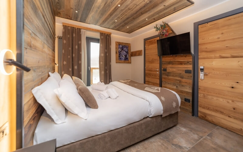 Cozy bedroom with a central bed, wooden accents, flat-screen TV, and natural light from a window with brown curtains.