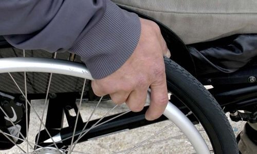 Una mano tiene una ruota di bicicletta o sedia a rotelle, con un sedile grigio e un pavimento in marmo sfocato.