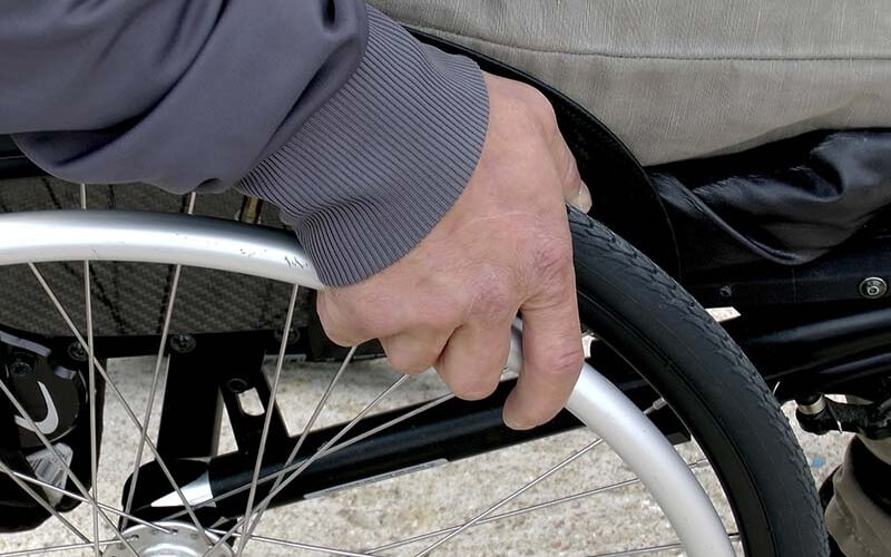 A hand grips the front wheel of a wheelchair on a light surface, with a gray jacket and a bag attached beneath the seat.