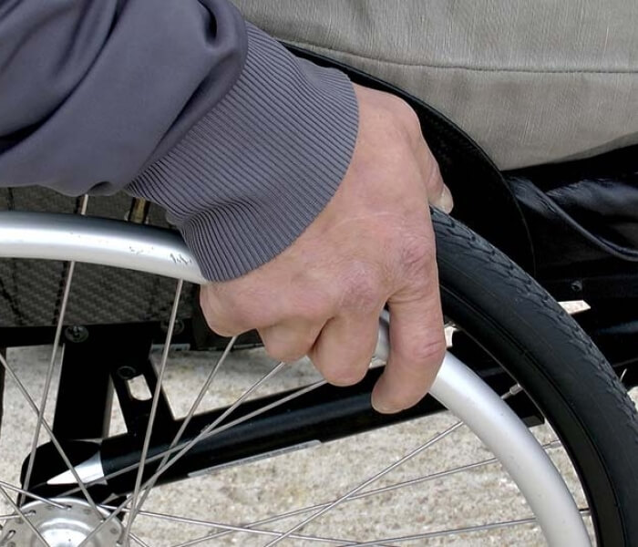 A hand in a gray shirt grips a wheelchair wheel, showing signs of aging, against a concrete surface, symbolizing determination.