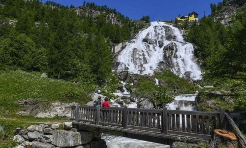 Rocky mountain waterfall with lush greenery, wooden walkway, and three individuals in vibrant attire under a sunny blue sky.