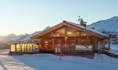 Ski lodge in snow-covered mountains at golden hour, with people on the roof and a warm ambiance contrasting the winter scene.