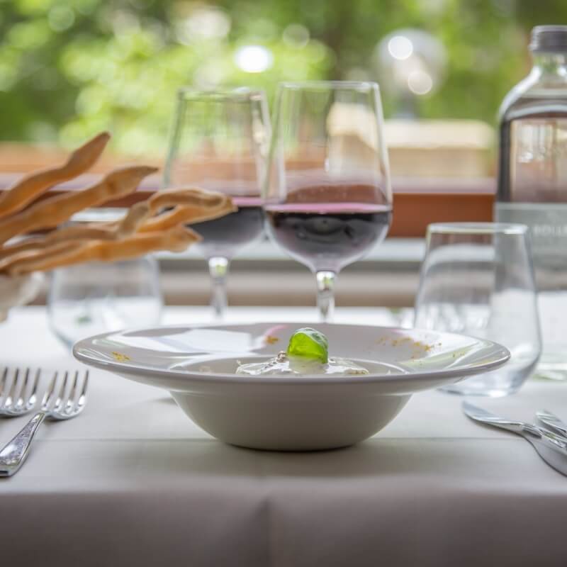 Formal table setting with a white bowl, wine glasses, silverware, and a blurred outdoor background with greenery.