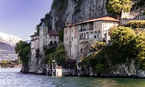 Historic multi-level structure on a cliff, overlooking water and mountains, with a weathered facade and lush greenery.