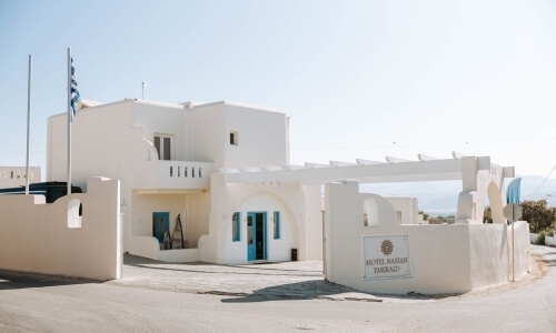 Edificio bianco con porte blu, bandiera greca, cancello e insegna "HOTEL MESSARI" sotto un cielo sereno e blu.