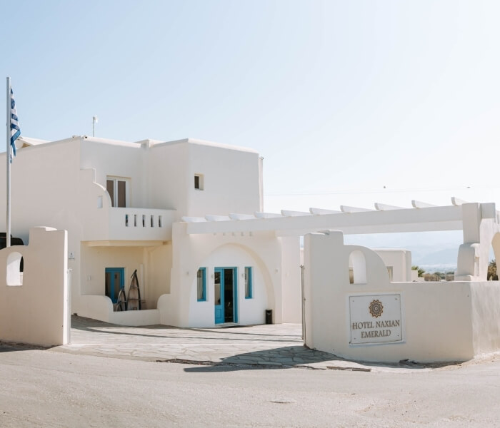 Edificio bianco con insegna "HOTEL NAXIAN EMERALD" in oro, bandiera greca, atmosfera serena e design elegante.