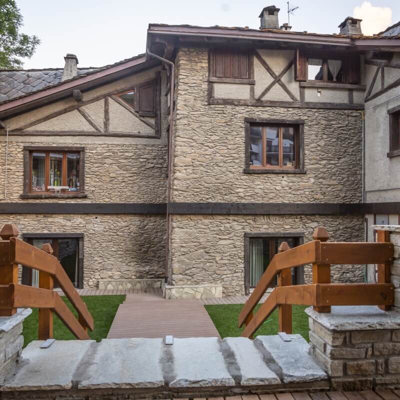 Casa in pietra a due piani con ponte di legno e recinzione, circondata da un'area erbosa.