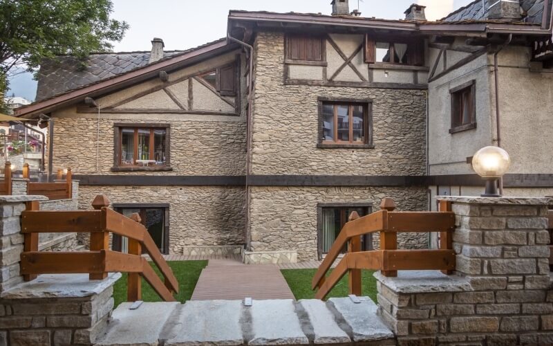 Two-story stone building with a brown roof, wooden bridge, grassy area, and gray overcast sky.