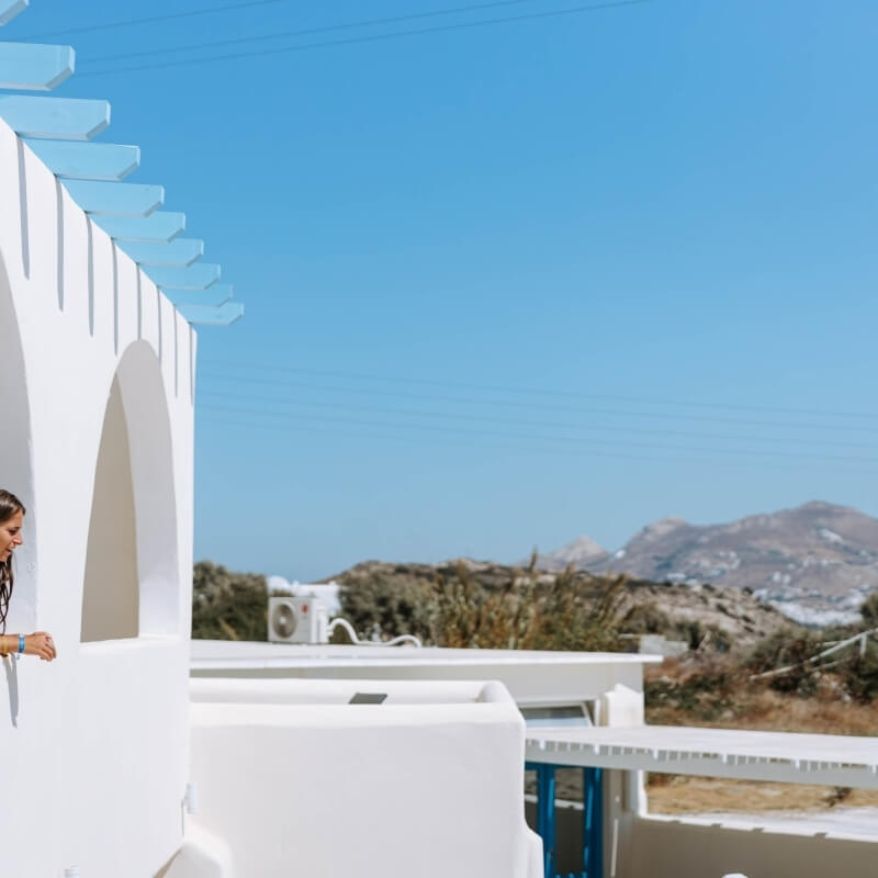 A woman in a white shirt stands in an arched doorway, overlooking a dry hillside and mountains under a clear blue sky.