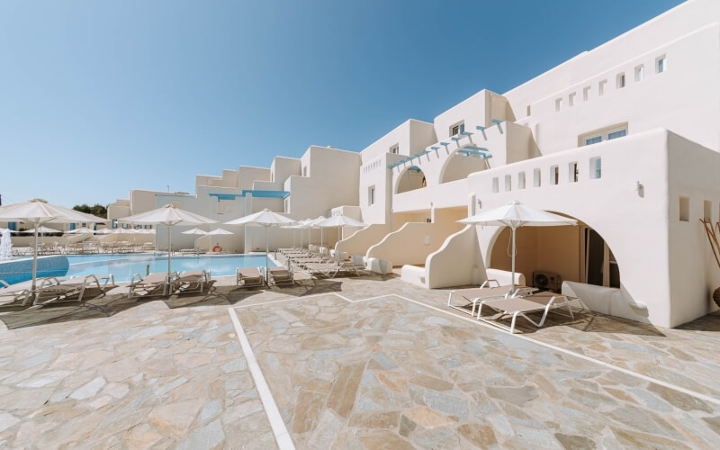 Serene outdoor pool area with white buildings, stone patio, lounge chairs, and bright blue water under a clear blue sky.