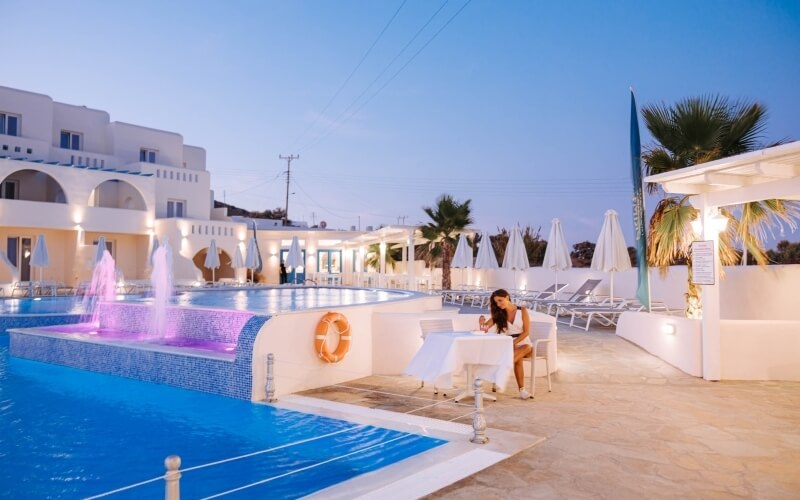 A woman in a white outfit sits by a serene pool with a pink fountain, surrounded by umbrellas and palm trees under a blue sky.
