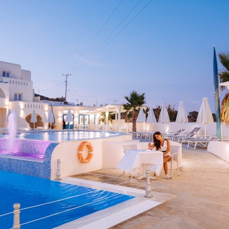 A woman sits at a table by a tranquil pool at dusk, surrounded by palm trees and a softly lit building.