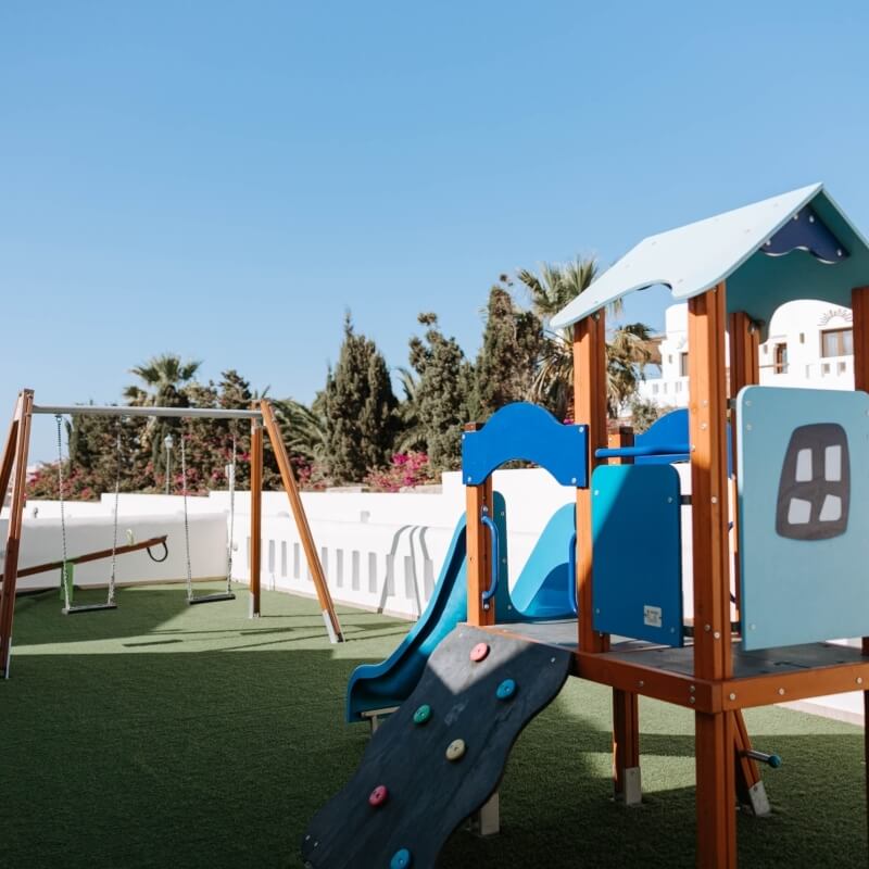 Colorful playground with swings, climbing structure, slide, and tunnel, set against trees and buildings under a clear blue sky.
