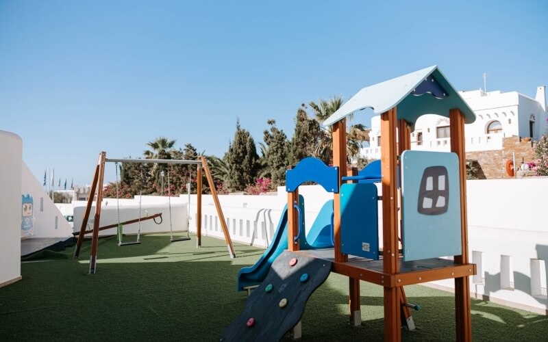 Vibrant playground with blue and brown play structure, slide, climbing wall, and rope ladder on green turf, white fence and building.