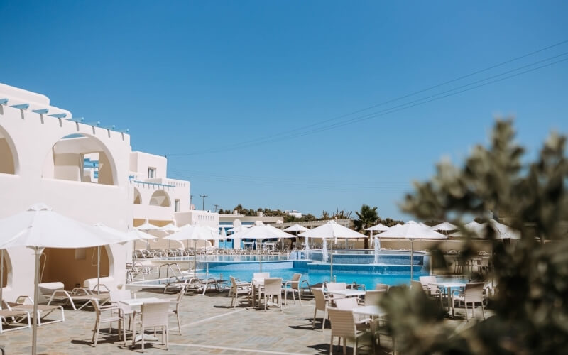 Serene pool area with white buildings, umbrellas, and a fountain, surrounded by greenery and a clear blue sky.