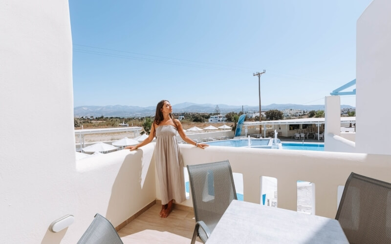 A woman in a light dress stands on a balcony, overlooking a pool and scenic landscape with rolling hills and a clear sky.