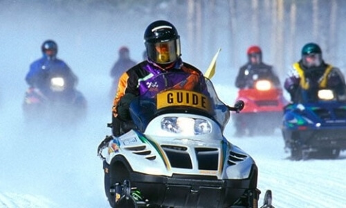 A snowmobile with a yellow "GUIDE" sign leads a group on a snowy trail, surrounded by snowy trees.